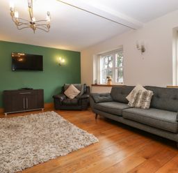 A living room with a green accent wall a mounted television a dark wood cabinet a dark leather armchair a gray sofa with cushions and a beige rug on wooden floor at About Time Cottage in Parkend