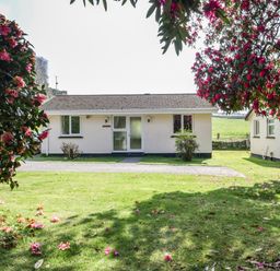 A single story house with white walls and a tiled roof surrounded by flowering trees and green grass at Watergate Bay in Liskeard