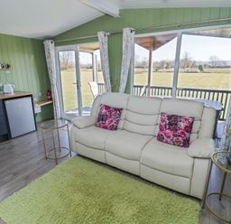 A living room with a white sofa with floral pillows green walls a small kitchenette and large windows at Cotswold View 1 in Honeybourne