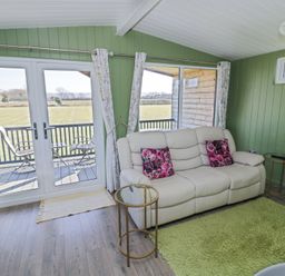 A living room with a cream sofa and glass tables next to windows overlooking a balcony and fields at Cotswold View 1 in Honeybourne