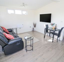 A living room with a black sofa with red pillows a glass coffee table a small dining table with two chairs a wall-mounted tv and light wood flooring at Cotswold View 2 in Honeybourne