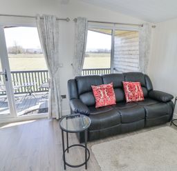 A living room with a black sofa with red pillows two round glass side tables and sliding glass doors leading to a balcony at Cotswold View 2 in Honeybourne