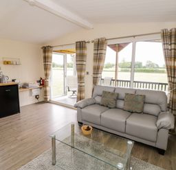A living room with a gray sofa and glass coffee table next to a kitchen area with a small fridge at Cotswold View 3 in Bretforton near Evesham