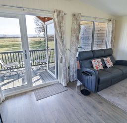 A living area with a dark sofa and patterned cushions near glass doors opening to a balcony with chairs at Cotswold View 4 Bretforton near Evesham