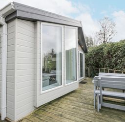 An outdoor wooden deck with a gray picnic table and a small house with large windows at Oyster Bay Lodge in Dalbeattie