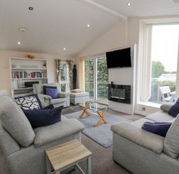 A living room with grey sofas and blue cushions a glass coffee table a wall mounted television and large windows at Oyster Bay Lodge in Dalbeattie