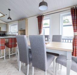 A dining area with a wooden table and grey chairs next to windows with red plaid curtains and a kitchen counter with red plaid bar stools at Beachcliff in Porth