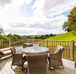 An outdoor wooden deck with a round table surrounded by wicker chairs and a lounge chair with green fields and trees in the background at Frogwell Chapel in Frogwell near Callington