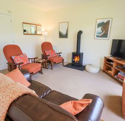 A living room with two armchairs a wood stove a television on a wooden stand and a leather sofa at Limhus Cottage in Keswick