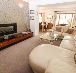 A living room with a beige sectional sofa a glass coffee table a mounted fireplace and a dining area at Fir Trees in Bowness-On-Windermere