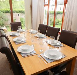 A dining room with a wooden table set with white plates bowls forks knives wine glasses and black chairs at Fir Trees in Bowness-On-Windermere