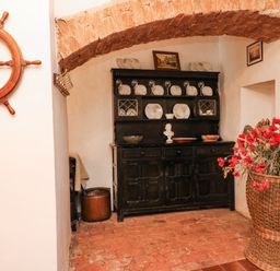A room with a wooden ship wheel on the wall a black cupboard with plates and glasses and a wicker basket with red flowers at Harbour Cottage in Watchet