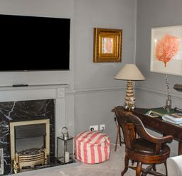 A corner room with a fireplace a wall-mounted TV a striped pouf a wooden desk with books two lamps and a wooden chair at Ferrystone in Kingswear near Dartmouth