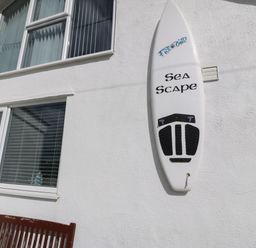 A white surfboard mounted on a white exterior wall next to two windows and a wooden bench at Sea Scape in Dinas Dinlle