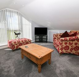 A sitting area with patterned armchairs a wooden coffee table a flat screen tv and large windows with vertical blinds at Sea Scape in Dinas Dinlle