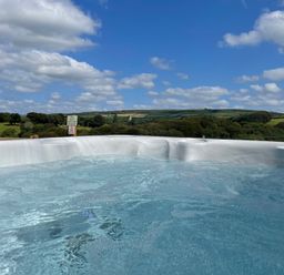 An outdoor hot tub with bubbling water overlooking a green landscape with trees and fields at Woodhall in Cardinham