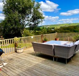 A wooden deck with rattan outdoor seating a table potted plants and a barbecue overlooking a green landscape at Woodhall in Cardinham