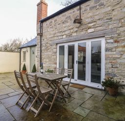 An outdoor patio area with a wooden table and four chairs next to stone wall with white doors and potted plants at Magnolia Barn in Mark