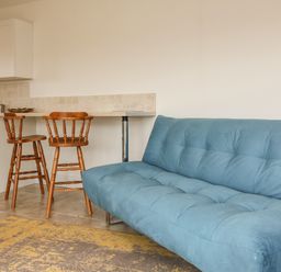 A kitchen area with white cabinets and a tiled backsplash with two wooden bar stools and a blue sofa with a patterned rug in front at The Stables in Crowcombe