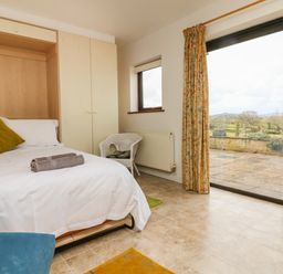 A bedroom with a bed white linens a green pillow towels on bed built in wardrobe a wicker chair tiled floor and sliding glass door overlooking a patio and countryside at The Stables in Crowcombe