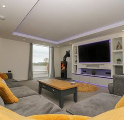 A living room with a gray sofa and yellow cushions a wooden coffee table a wall-mounted television and a wood-burning stove at Valley View in Mullion