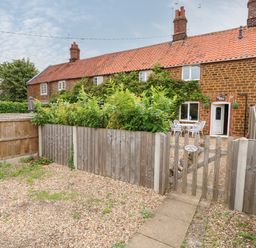 A fenced garden with gravel and a wooden gate in front of a brick house with red roof tiles at Tawny Cottage in Heacham