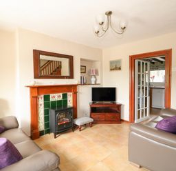 A living room with two sofas facing each other a fireplace with green tiles and a small stove a wooden TV stand with a TV and a small upholstered footstool at Tawny Cottage in Heacham
