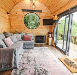 A wooden room with a grey sectional sofa and cushions a circular window a wall-mounted tv and a glass door leading outside at Offas Dyke Escape in St Asaph