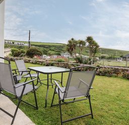 An outdoor patio with a table and four chairs on grass with bushes hills and houses in the background at 7 Europa Court in Mawgan Porth