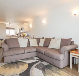 A living room with a brown corner sofa and a rug with circular patterns adjacent to a kitchen at 7 Europa Court in Mawgan Porth