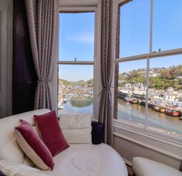A seating area with white and purple cushions next to large windows overlooking a harbor with boats and hillside houses at Albany in Looe