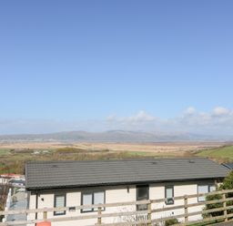 A house with a grey roof and multiple windows with a wooden fence in front and a landscape of fields and hills under a blue sky at McAuliffe Lodge in Borth
