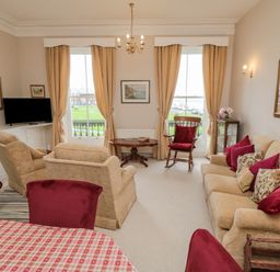 A living room with beige sofas and armchairs red cushions and curtains and a dining table with red chairs at Sandsend Sunset View Whitby