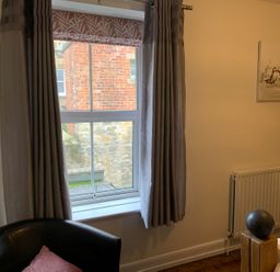 A room with a window showing a brick wall outside gray curtains a black chair with a cushion and a radiator with a painting of birds on the wall at A Stone's Throw in Alnmouth