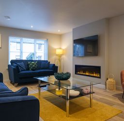 A living room with blue sofas a glass coffee table a yellow rug a wall-mounted television and a fireplace at The Neon Rose in Bowness-On-Windermere