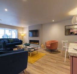 A living room with blue sofas a glass coffee table a wall-mounted TV an electric fireplace a round pink chair and a dining area with white chairs and a marble countertop at The Neon Rose in Bowness-On-Windermere