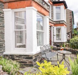 A small outdoor patio with a metal bench and table holding two glasses in front of a stone house with white framed windows at Oakhurst in Keswick