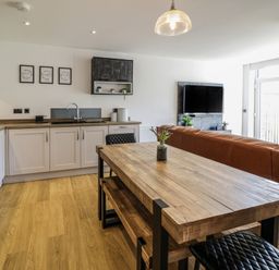 An open plan kitchen and living area with a wooden dining table black chairs a brown sofa a wall-mounted TV and kitchen cabinets at 2 Pensyflog Barns in Porthmadog