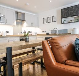 An open kitchen and dining area with wooden table, black chairs, white cabinets, and a brown leather sofa with cushions at 2 Pensyflog Barns in Porthmadog