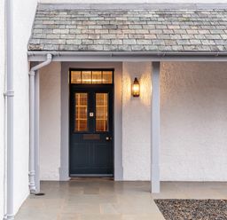 A dark green front door with glass panels under a small covered porch with a lit wall lantern at The Vicarage near Rosthwaite
