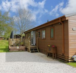 A wooden cabin with a small porch and gravel driveway surrounded by grass and trees at Lakeland Dream @ South Lakeland Leisure Lodge in Warton
