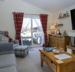 A living room with a plaid armchair and ottoman a wooden coffee table a wooden sideboard with lamps and decor and a wall-mounted TV at Lakeland Dream @ South Lakeland Leisure Lodge in Warton