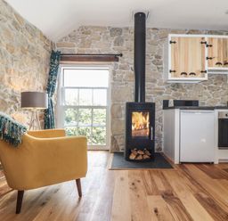 A room with stone walls a yellow armchair a floor lamp a stove with fire wooden cabinets and a window at The Hayloft in Helston