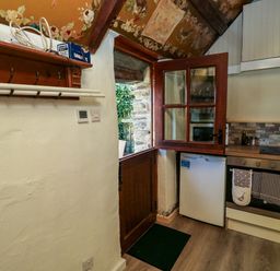 A kitchen with a wooden door open to the outside a small fridge an oven with towels a countertop with kitchen utensils and a shelf with hooks at Toad Hall Cottage in Hutcherleigh near Blackawton