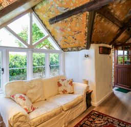 A room with a beige sofa and floral cushions by a window with patterned orange ceiling and wooden beams at Toad Hall Cottage in Hutcherleigh near Blackawton