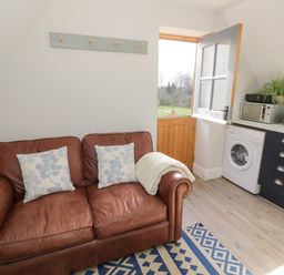 A living area with a brown leather sofa with cushions next to an open door leading outside and a kitchen area with appliances at Coach House Mews in Alderminster