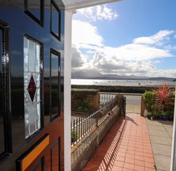 View from a black front door opening to a tiled pathway with metal fence and plants overlooking a road and water with boats and mountains in the distance at Lluest in Beaumaris