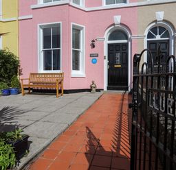 An entrance area with a black gate and a pathway leading to black doors of a pastel pink and beige building with a wooden bench and plants at Lluest in Beaumaris