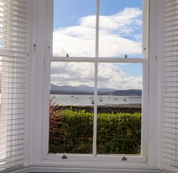 A window with white blinds showing a view of boats on water and mountains in the distance at Lluest in Beaumaris