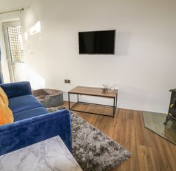 A living room with a blue sofa and yellow cushions a mounted TV a wood stove a dog bed and a small table at Primrose End in Jacobstow